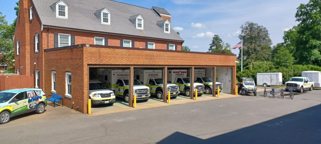 A view of a brick building serving as a fire station with multiple emergency vehicles, including vans and a firetruck, parked in and around a three-bay garage under a clear blue sky.