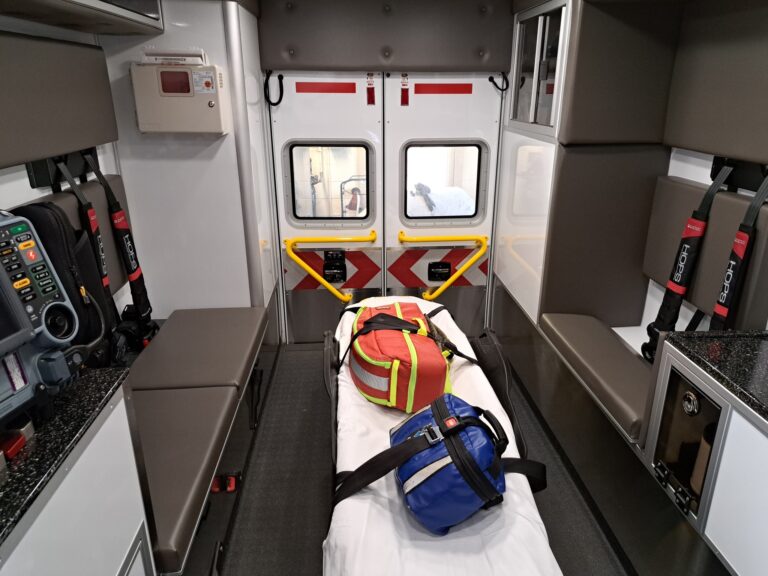 Interior of an ambulance showing a stretcher with a yellow and red bag, medical equipment, and seating along the sides.