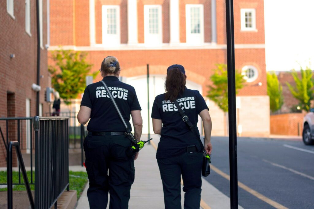 Two rescue workers wearing matching "Fredericksburg Rescue" shirts walk side by side on a paved walkway. They carry emergency gear, and a brick building with white-trimmed windows is visible in the background. The scene is well-lit with natural daylight.