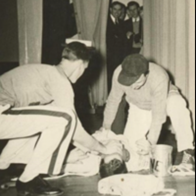 Two individuals, one in a baseball cap, are attending to a person lying on the ground in what appears to be an indoor setting. Two more individuals watch from behind a curtain in the background. A bucket and some cloth are on the floor beside them.