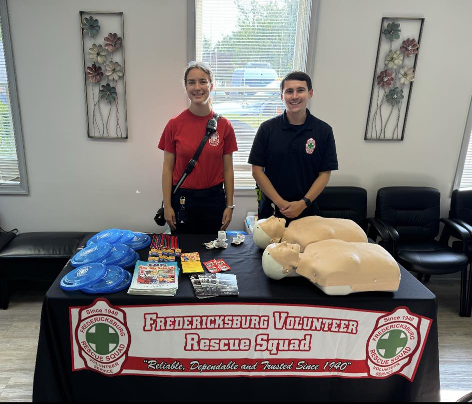 Two Fredericksburg Volunteer Rescue Squad members, a woman on the left and a man on the right, stand behind a table with CPR mannequins, pamphlets, frisbees, and keychains. The table displays the squad's name and logo. Walls are adorned with floral artwork.