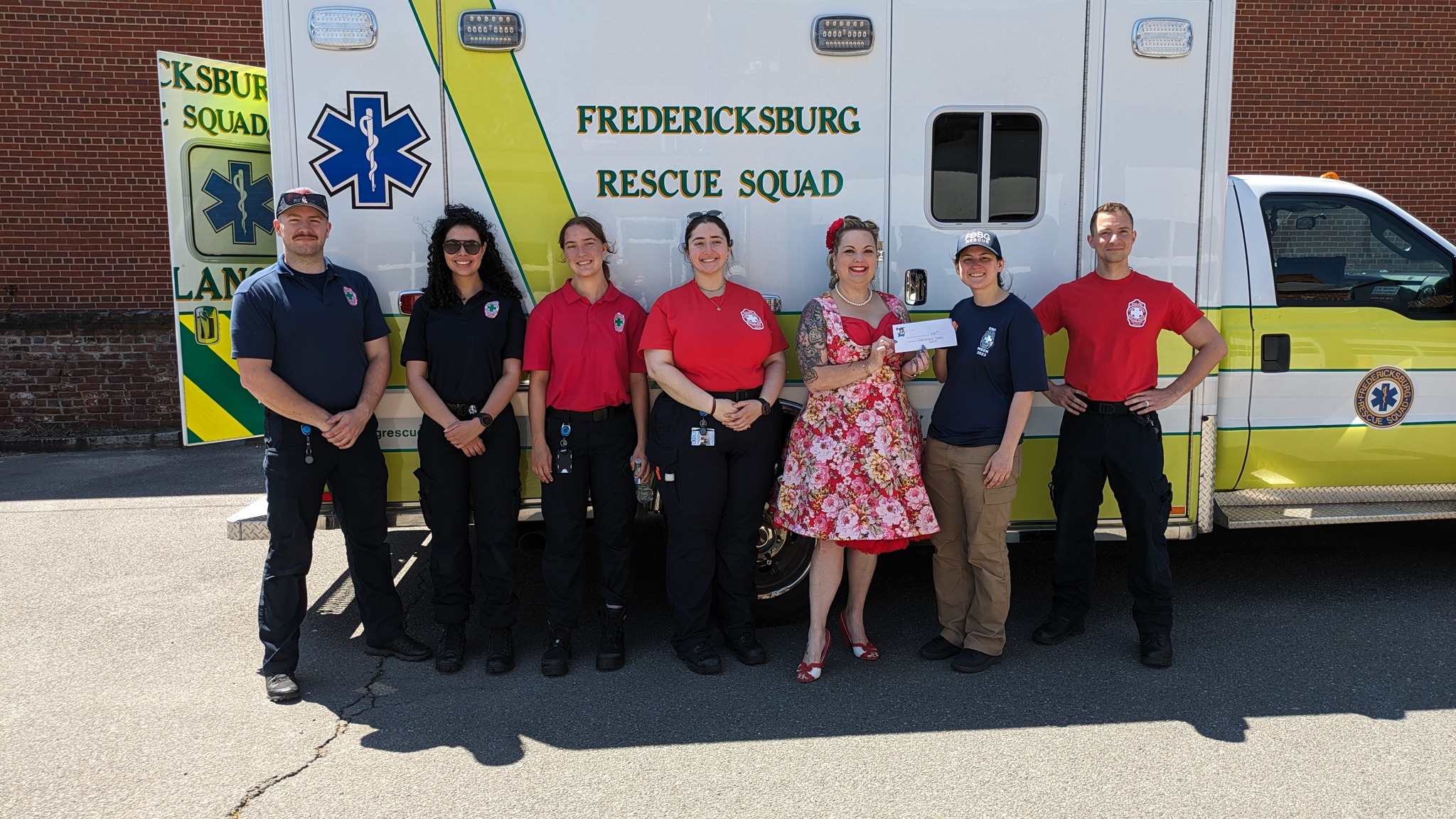 A group of seven people stands in front of an ambulance with "Fredericksburg Rescue Squad" written on it. Five of them are wearing rescue squad uniforms, and two are in casual attire, one holding a certificate. They are outdoors on a sunny day.