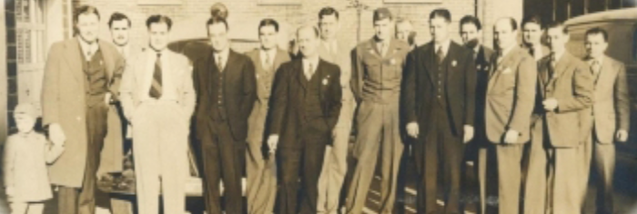 A sepia-toned vintage photograph shows a group of men in suits and military uniforms standing side by side. The men form a straight line and are outdoors in front of a brick building, accompanied by a young child holding a man's hand to the left of the group.