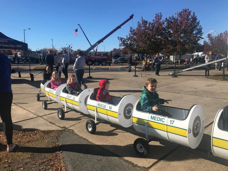 Several children are riding in individual train-like cars designed to resemble rescue vehicles on a sunny day at an outdoor event. A fire truck and crane are visible in the background, along with an American flag and early autumn trees. Adults stand nearby.