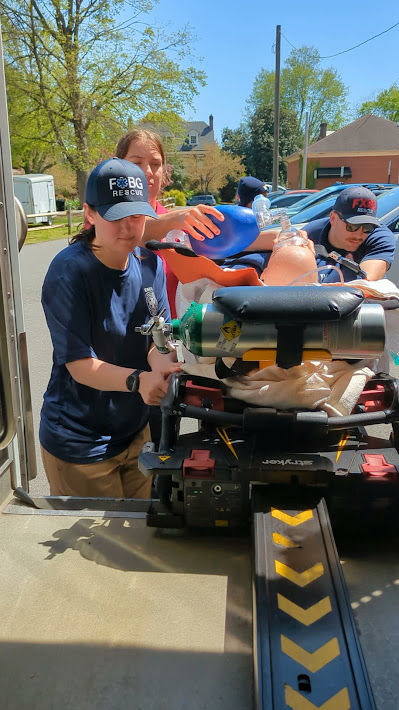 PXL_20240414_180053649.TS~2 Paramedics carefully load a stretcher with medical equipment and a mannequin into an ambulance. One paramedic secures the gear while another guides the stretcher. They are wearing navy blue shirts and caps with "FBOG RESCUE" written on them. It is a sunny day.