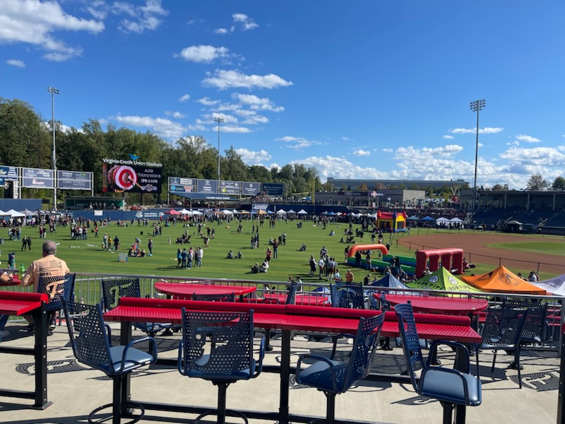 A sunny day at an outdoor event held in a baseball stadium. People are scattered across the field, some near tents and inflatables. Empty seating with red countertops is in the foreground. The sky is clear with scattered clouds. Trees line the background.